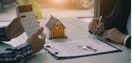 Αγοραστής υπογράφει συμβόλαιο ενώ πωλητής του παρουσιάζει τιμή σε αριθμομηχανή. Δίπλα στο συμβόλαιο υπάρχει μοντέλο σπιτιού με τα κλειδιά επάνω