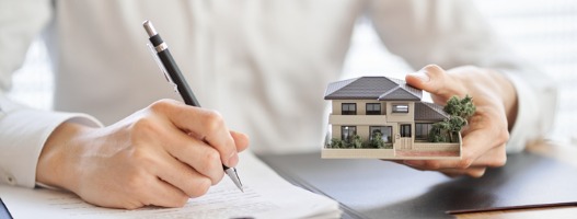 Άνθρωπος υπογράφει συμβόλαιο ενώ κρατά στο ένα χέρι μοντέλο σπιτιού
